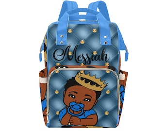 May include: Blue diaper bag with a quilted design and gold accents. The bag features a cartoon image of a Black baby boy wearing a crown and holding a blue dummy. The name "Messiah" is printed on the front of the bag.