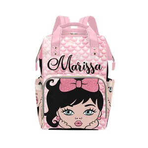 Puede incluir: Mochila rosa con un patrón de escamas de sirena y el nombre "Marissa" en escritura negra. La parte delantera presenta una niña de dibujos animados con un lazo rosa, cabello negro y ojos azules. Incluye bolsillos laterales y asas superiores.