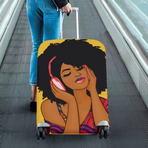 May include: A yellow suitcase featuring a stylized illustration of a woman with closed eyes wearing pink headphones. The woman has dark skin, black curly hair, and is wearing a patterned top. The suitcase has a telescoping handle and black wheels.