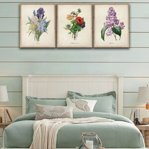May include: Three framed botanical prints featuring a blue iris, a red and white anemone, and a purple lilac. The prints are hung on a light blue wall above a white bed with a light green duvet and a white throw blanket.
