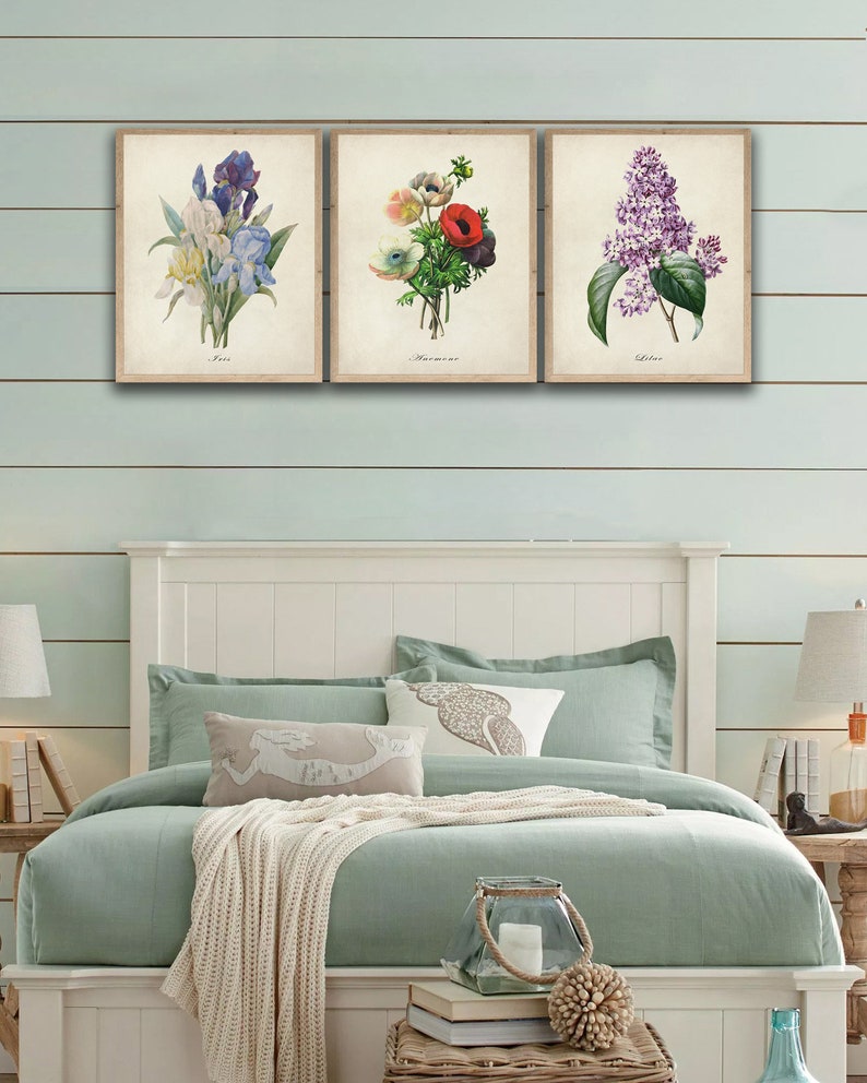 May include: Three framed botanical prints featuring a blue iris, a red and white anemone, and a purple lilac. The prints are hung on a light blue wall above a white bed with a light green duvet and a white throw blanket.
