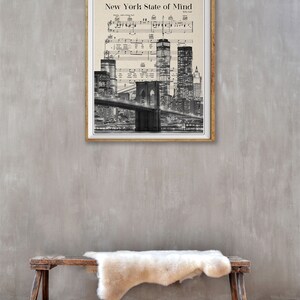 May include: A black and white print of the New York City skyline with the Brooklyn Bridge in the foreground. The print is framed in a wooden frame and has the song title "New York State of Mind" and the songwriter's name "Billy Joel" printed at the top.