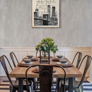 May include: A black and white print of the New York City skyline with the Brooklyn Bridge in the foreground. The print is framed and hanging on a gray wall above a wooden dining table with black metal chairs.