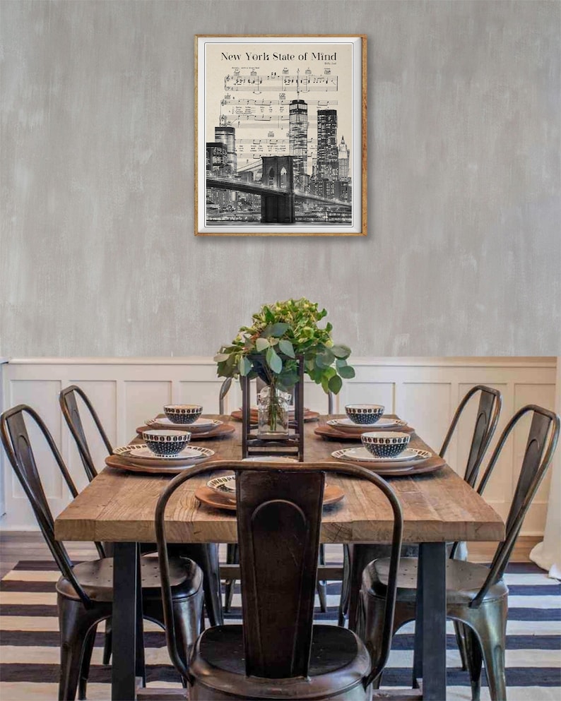 May include: A black and white print of the New York City skyline with the Brooklyn Bridge in the foreground. The print is framed and hanging on a gray wall above a wooden dining table with black metal chairs.