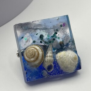 May include: A square resin ring featuring a blue and silver design, with three seashells and glitter inclusions. The ring has a silver-coloured band.