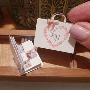 Può includere: Un piccolo sacchetto regalo bianco con un nastro rosa e le iniziali "F & M" in un design floreale a forma di cuore. Il sacchetto è appoggiato su una superficie di legno accanto a un piccolo album fotografico.