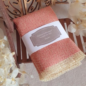 Peut inclure: Une couverture miniature pliée de couleur corail avec un tissage texturé et des franges couleur crème aux extrémités. Une bande de papier blanc entoure le centre, portant le texte imprimé : Fashion in Miniature, Snuggle me tight, Blanket.