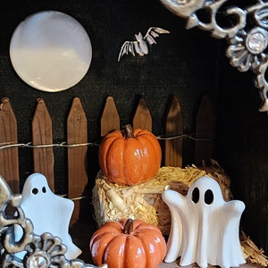 May include: A miniature Halloween scene with a white moon, a bat, a brown picket fence, two white ghosts, and two orange pumpkins on a bed of straw.