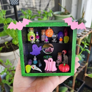 May include: A green wooden box with a black interior, decorated with pink bat wings, contains a miniature Halloween-themed scene. The scene includes potion bottles, a ghost, a skull, and other spooky decorations.