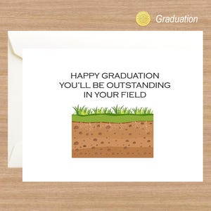 Op de afbeelding: Een witte kaart met de tekst "Happy Graduation You'll Be Outstanding In Your Field" en een cartoon afbeelding van een stukje gras en aarde.