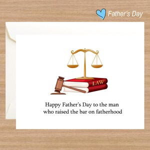 May include: A white card with a gold scale of justice, a gavel, and a stack of books with the word "LAW" on the cover. The card has the text "Happy Father's Day to the man who raised the bar on fatherhood."