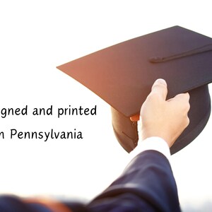 Op de afbeelding: Een zwarte graduatiecap met kwast wordt vastgehouden door een hand in een donkerblauw pak. De tekst "Designed and printed in Pennsylvania" is zichtbaar in de afbeelding.