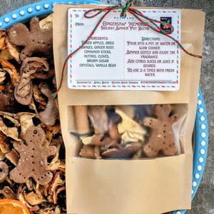 May include: A brown paper pouch labeled Gingersnap Memories Holiday Simmer Pot Blend containing dried apples, orange slices, cinnamon sticks, cloves, and gingerbread man-shaped pieces, tied with red and green twine, resting on a blue-rimmed bowl.