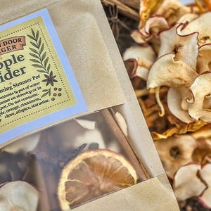 以下が含まれることがあります： 「Kitchen Door Forager Apple Cider」と書かれたパッケージのクローズアップ。透明な窓から乾燥オレンジスライス、シナモンスティック、リンゴスライスが見える。背景には乾燥リンゴスライス。ラベルには使用方法が記載されています。