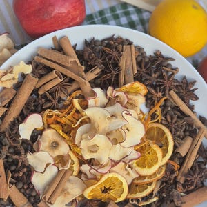 May include: A white bowl filled with a mixture of dried apple slices, dehydrated orange slices, whole star anise, cinnamon sticks, and dark peppercorns. A brown paper pouch labeled Apple Cider sits behind the bowl with a fresh apple and orange.