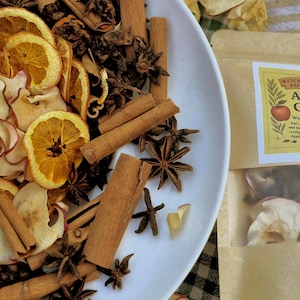 May include: A white plate filled with dried apple and orange slices, cinnamon sticks, and star anise. A package of "Apple Cider" warming simmer pot ingredients is visible on the right.