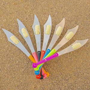 May include: A set of eight colorful corn cob holders with a silver finish. The holders are arranged in a fan shape with the handles pointing outwards. Each holder has a small, yellow corn cob inside.