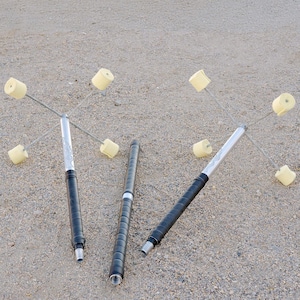 May include: Four black and silver fire staffs with yellow fire poi attached. The staffs are laid out on a sandy surface.