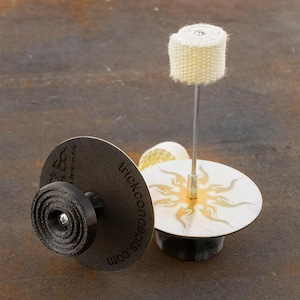 May include: Two metal fire starters with a sun design. One fire starter has a black base and a white fire starter on a wire.