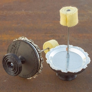 May include: Two silver-colored metal candle holders with black bases. Each holder has a decorative, scalloped edge. One holder has a yellow wick on a metal rod. The other holder has a black base with the text "trickconcepts.com" and "Wickcepts Locks Better on Fire!" printed on it.