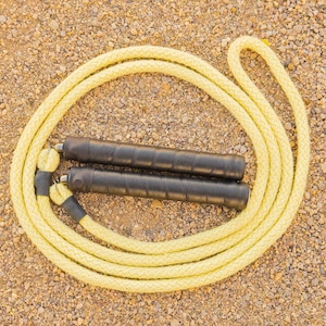 May include: A yellow jump rope with black handles lies on a textured, tan surface. The rope is coiled, showing its woven texture. The handles are cylindrical and appear to be wrapped in a textured material.