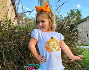 May include: A white short-sleeved shirt with a pumpkin design and the letters "SSM" embroidered on the pumpkin. The shirt is paired with yellow and white striped pants with a ruffle at the bottom.