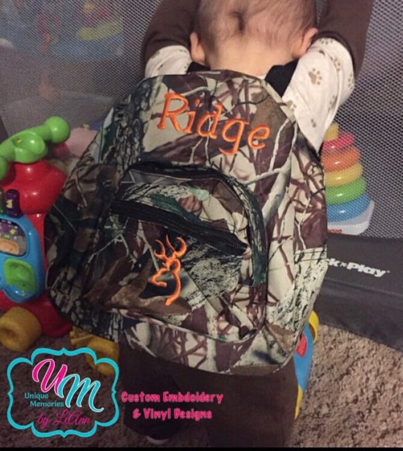 May include: A camouflage backpack with a brown, orange, and green pattern. The backpack has a zippered pocket and a buckled strap. The name "Ridge" is embroidered on the front of the backpack in brown thread. A buck logo is embroidered on the front of the backpack in orange thread.