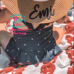 May include: A straw sun hat with a white band and the name "Emi" embroidered in black. The hat is worn with a navy blue swimsuit with white heart patterns and red trim. The swimsuit has a ruffled skirt.