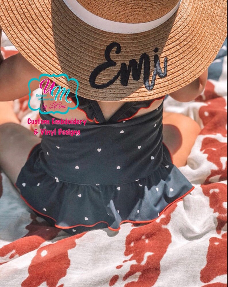 May include: A straw sun hat with a white band and the name "Emi" embroidered in black. The hat is worn with a navy blue swimsuit with white heart patterns and red trim. The swimsuit has a ruffled skirt.