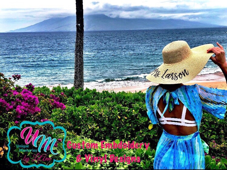 May include: A straw sun hat with the text "Mrs. Larson" embroidered on the brim. The hat is worn by a person standing on a beach with a view of the ocean and mountains in the background.