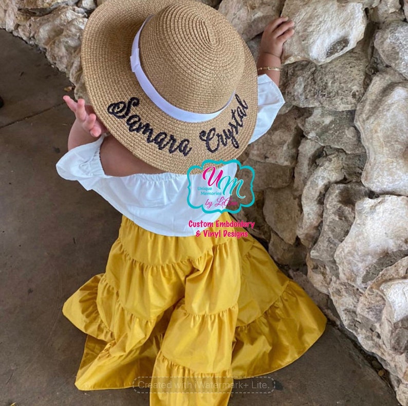 May include: A straw sun hat with a white band and embroidered text reading "Samara Crystal." The hat is paired with a white off-the-shoulder top and yellow tiered trousers. The image also includes a logo that says "Unique Memories by Lela."