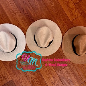 May include: Three straw sun hats in cream, tan, and brown, each with a black band. The hats are arranged on a wooden surface. The image includes a logo that says "Unique Memories" and "Custom Embroidery & Vinyl Designs."