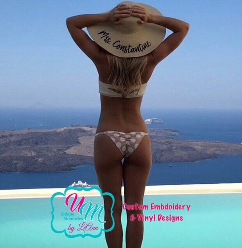 May include: A woman wearing a white bikini top and a polka dot bikini bottom is standing by a pool. She is wearing a large sun hat with the text "Mrs. Constantine" written on it. The image is taken from behind the woman, and the background is a blue sky and ocean.