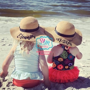 May include: Two children wearing straw sun hats with their names, "Eleanor" and "Violet", embroidered on the black ribbon bands. The children are sitting on a sandy beach.