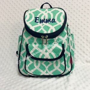 May include: A green and white quilted backpack with a black zipper and trim. The backpack has a pocket on the front and the name "Emma" embroidered on the top flap.