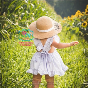 May include: A child in a light purple dress with frilled sleeves and a bow at the back, wearing a wide-brimmed straw hat with a white band. The hat has text on it. The child is in a field of tall green grass.
