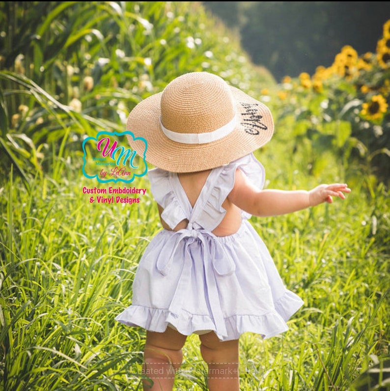 May include: A child in a light purple dress with frilled sleeves and a bow at the back, wearing a wide-brimmed straw hat with a white band. The hat has text on it. The child is in a field of tall green grass.