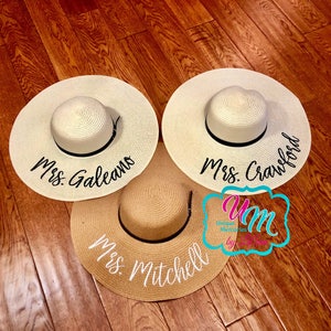 May include: Three straw sun hats with black ribbon trim and personalized embroidery. The hats are on a wooden floor. The text on the hats reads "Mrs. Galeano", "Mrs. Crawford", and "Mrs. Mitchell".