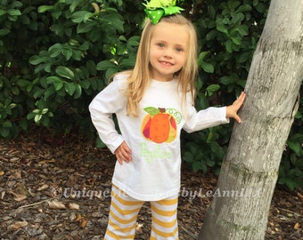 May include: A young girl wearing a white long-sleeved shirt with a pumpkin applique and the name "Peyton" embroidered on it. She is also wearing yellow and white striped pants with a ruffle at the bottom. She is standing next to a tree and has a green bow in her hair.