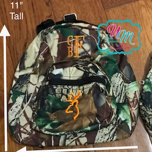 May include: A camouflage-patterned mini backpack with a black zipper and a brown and orange embroidered deer logo. The backpack is 11 inches tall and 9.5 inches wide.