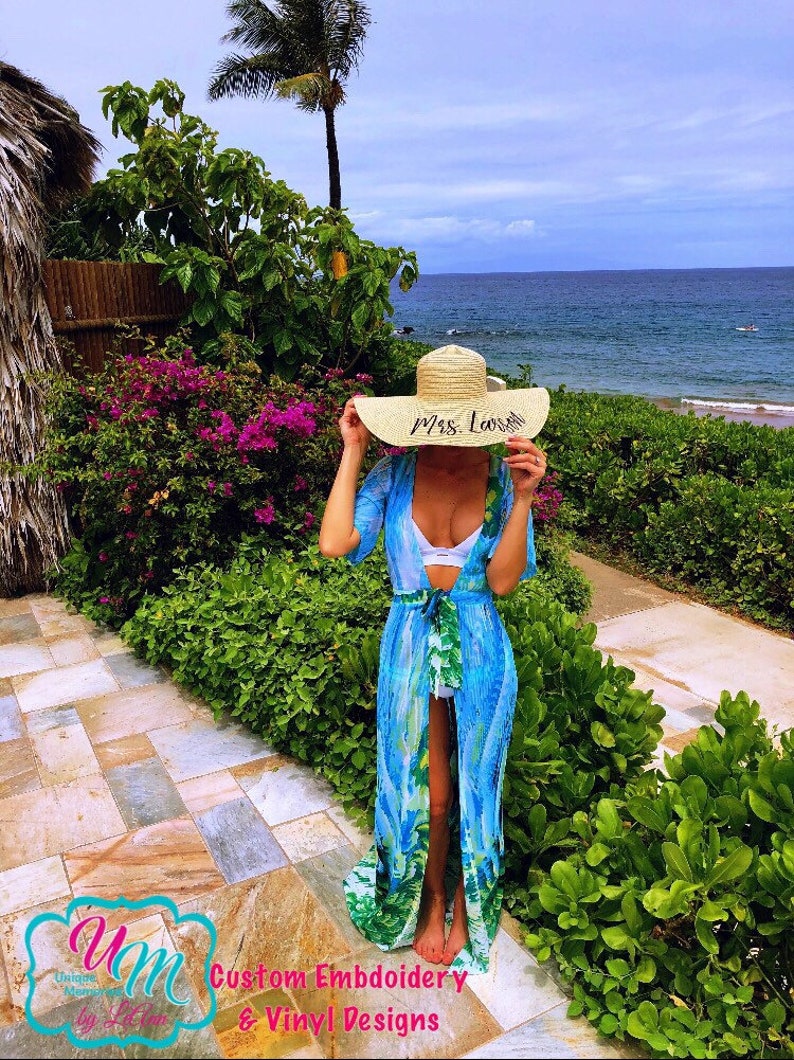 May include: A woman wearing a blue and green beach cover-up with a white bikini top underneath. She is holding a large straw sun hat with the text "Mrs. Lovell" embroidered on it. The woman is standing on a stone path with lush green foliage and a view of the ocean in the background.