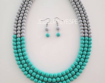 May include: A two-piece jewelry set featuring a layered necklace and matching earrings. The necklace is made of gray and turquoise beads, while the earrings are turquoise.