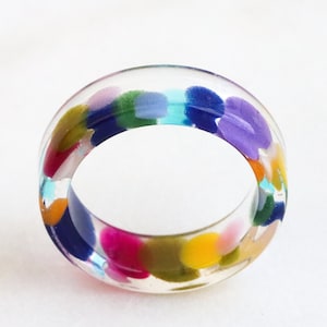 May include: A clear, circular ring made of resin, featuring a variety of embedded colorful spots in shades of blue, purple, yellow, pink, and green. The band has a smooth, polished finish and a rounded profile, showcasing the vibrant confetti-like pattern.