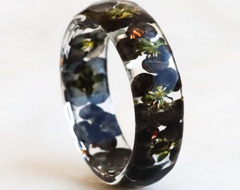 May include: A clear resin ring featuring embedded dark blue and black pressed flowers with small green accents. The band has a smooth, polished finish and a rounded profile, showcasing the botanical elements suspended within the transparent material.