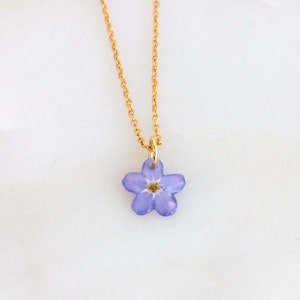 Puede incluir: Un delicado collar de cadena dorada con un pequeño colgante de flor de color lila claro. La flor está prensada y conservada en resina, lo que le da una apariencia translúcida.