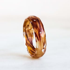 May include: A clear resin ring with a botanical design. The ring features a band of dried flowers in shades of brown, orange, and yellow. The ring is a unique piece of jewelry.