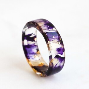 Puede incluir: Un anillo de resina transparente con detalles en morado y dorado. El anillo tiene una banda ancha y está decorado con remolinos de morado y dorado.