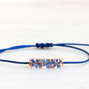 May include: A blue adjustable cord bracelet with a rectangular charm featuring pressed blue flowers.