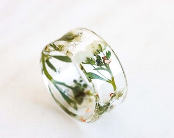 May include: A clear resin ring with white flowers and green leaves pressed inside. The ring is thick and has a rounded shape.