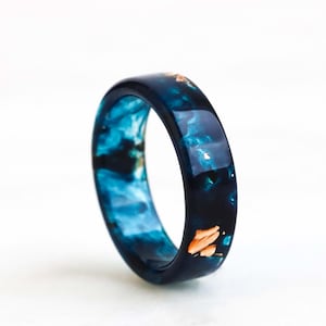 May include: A polished resin ring featuring a deep blue and black marbled pattern with embedded metallic copper-colored flakes. The band has a smooth, glossy finish and a rounded profile, showcasing a vibrant contrast between the dark tones and bright accents.